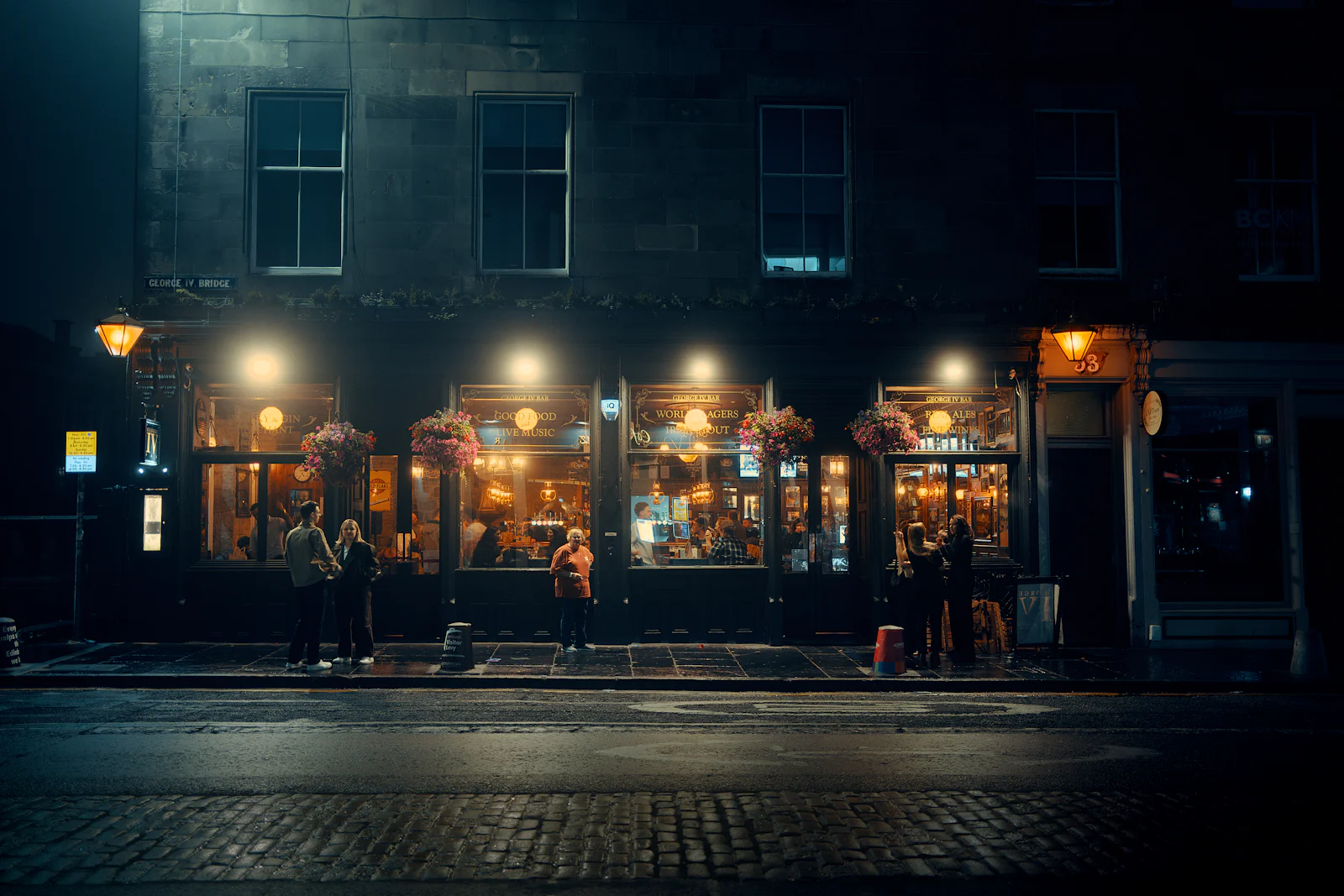 People outside a bar on an Edinburgh street at night, its windows lit warm under dark stone